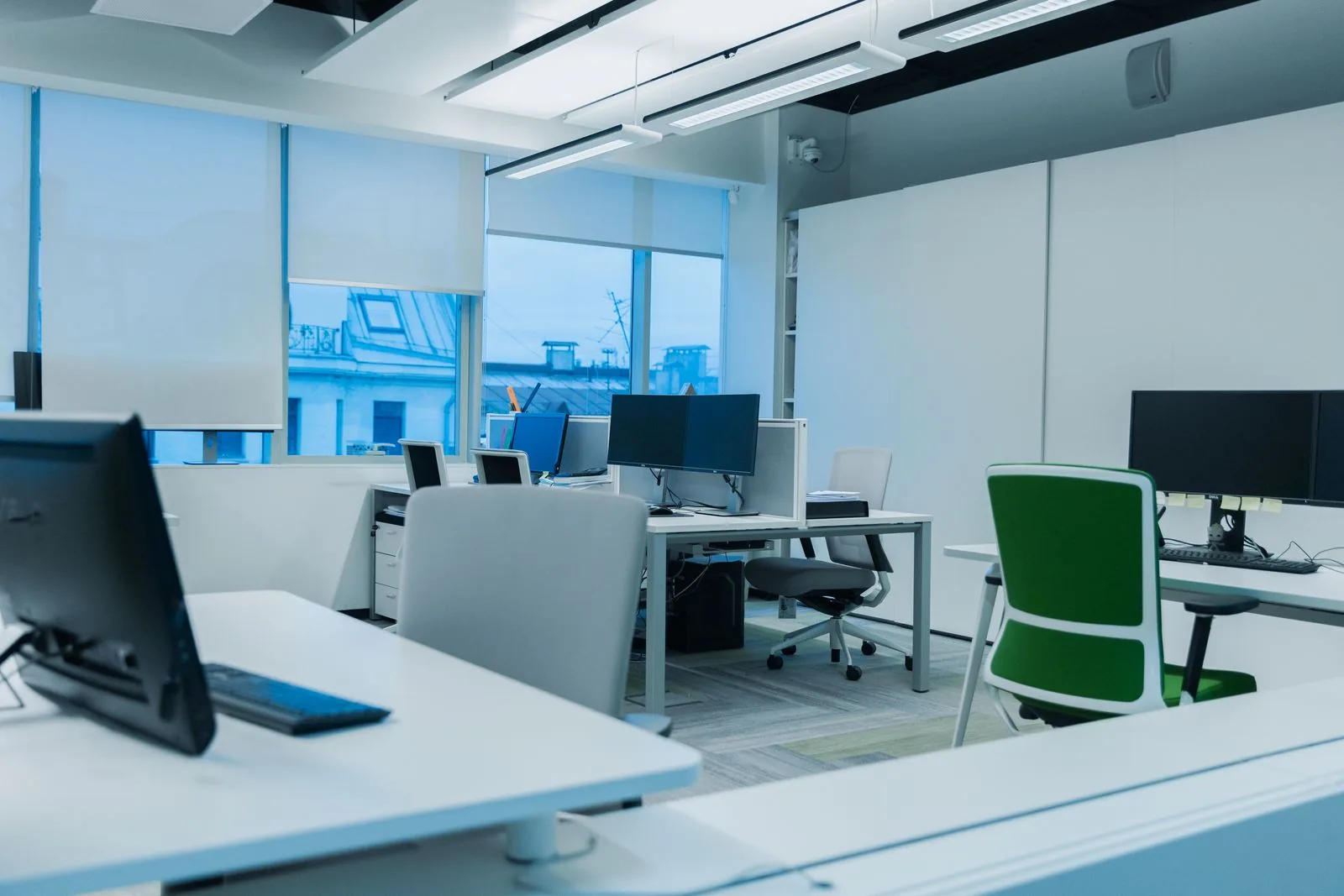 Was kostet Büroreinigung pro Quadratmeter? Helles Büro mit weißen Schreibtischen, Monitoren, Bürostühlen und einer Fensterfront mit Blick auf Nachbardächer