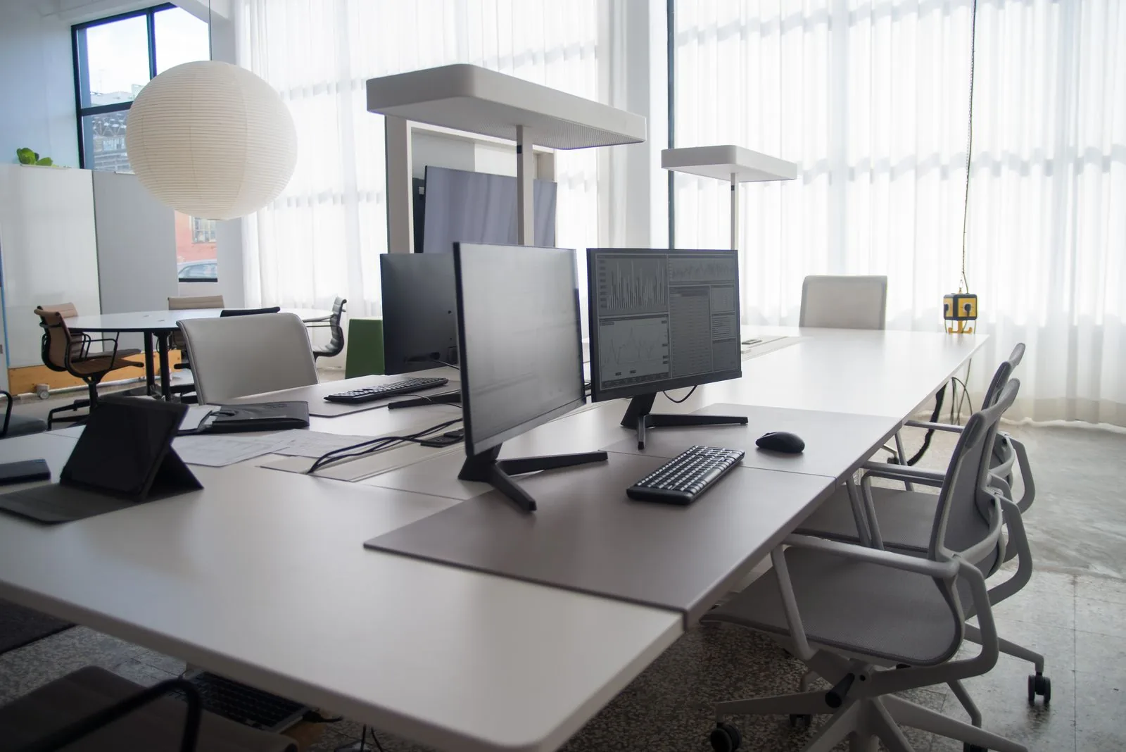 Wie oft sollte ein Büro gereinigt werden? Lange Schreibtischreihe in einem hellen Großraumbüro mit Monitoren, Bürostühlen und einer Fensterfront mit weißen Vorhängen