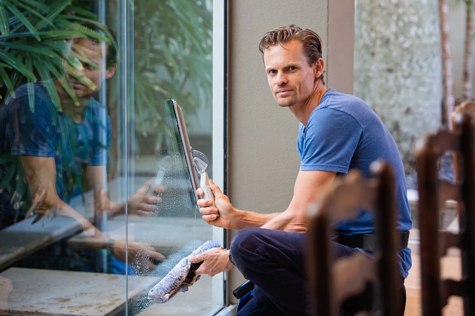 Wie oft sollten Fenster im Objekt gereinigt werden? Mann im blauen T-Shirt kniet an einer bodentiefen Glasscheibe und zieht sie mit einem Abzieher ab, in der Hand einen Einwascher