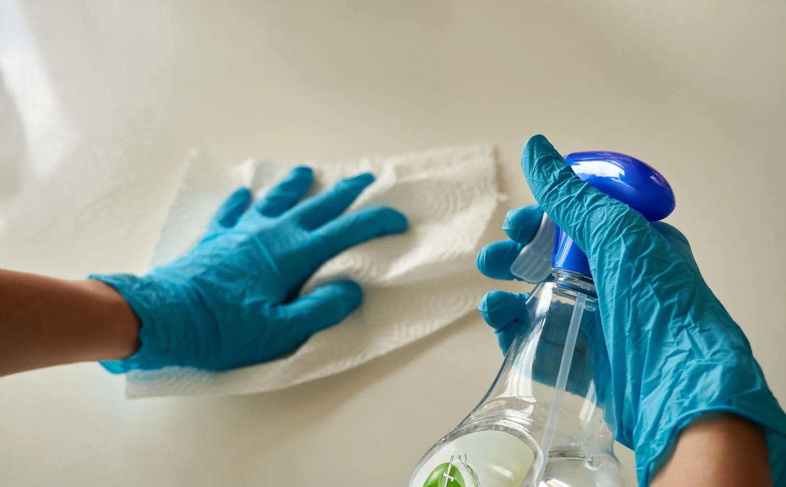 Flächendesinfektion: warum die Einwirkzeit über alles entscheidet Zwei Hände in blauen Einweghandschuhen wischen eine helle Fläche mit einem Papiertuch, daneben eine Sprühflasche