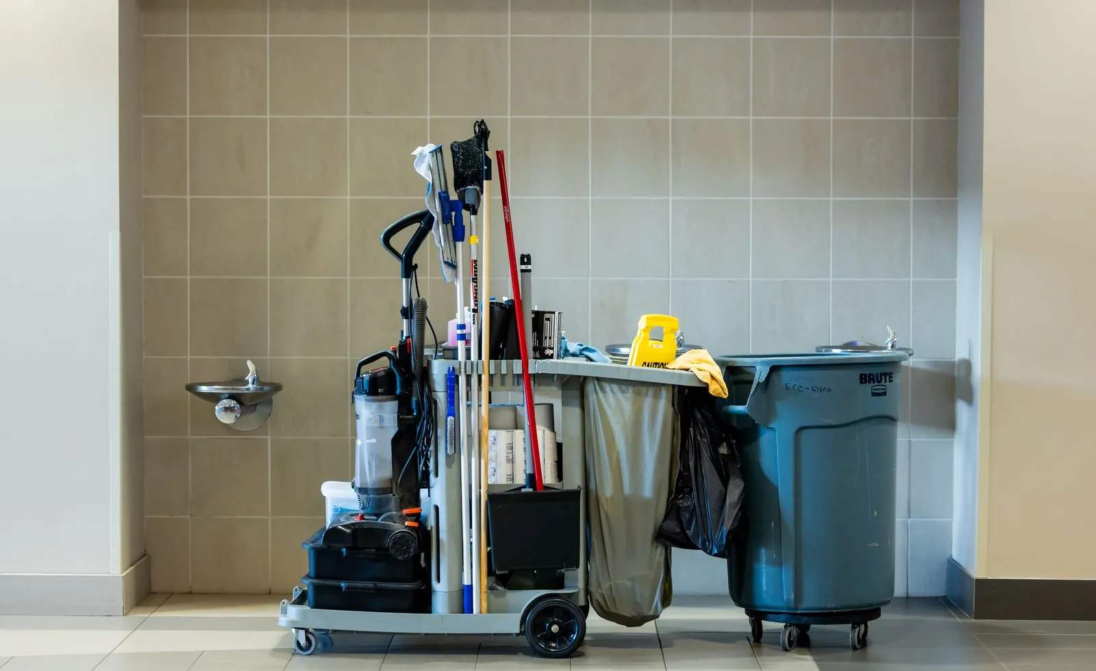 Grundreinigung oder Unterhaltsreinigung: der Unterschied Voll bestückter Reinigungswagen mit Wischmopps, Staubsauger, Müllsack und Warnschild vor einer grau gefliesten Wand