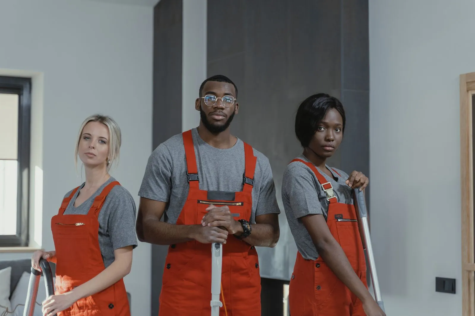 Woran Sie eine gute Reinigungsfirma erkennen Drei Reinigungskräfte in roten Latzhosen und grauen Shirts stehen mit Staubsauger und Wischgerät in einer hellen Wohnung