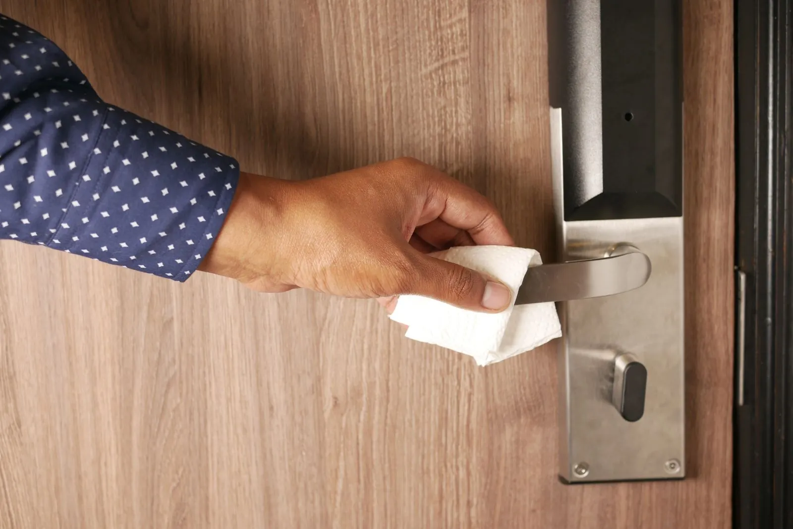 Kontaktflächen richtig reinigen: die unterschätzte Liste Hand wischt die Klinke einer Holztür mit einem weißen Papiertuch ab, daneben das Türschloss aus Edelstahl