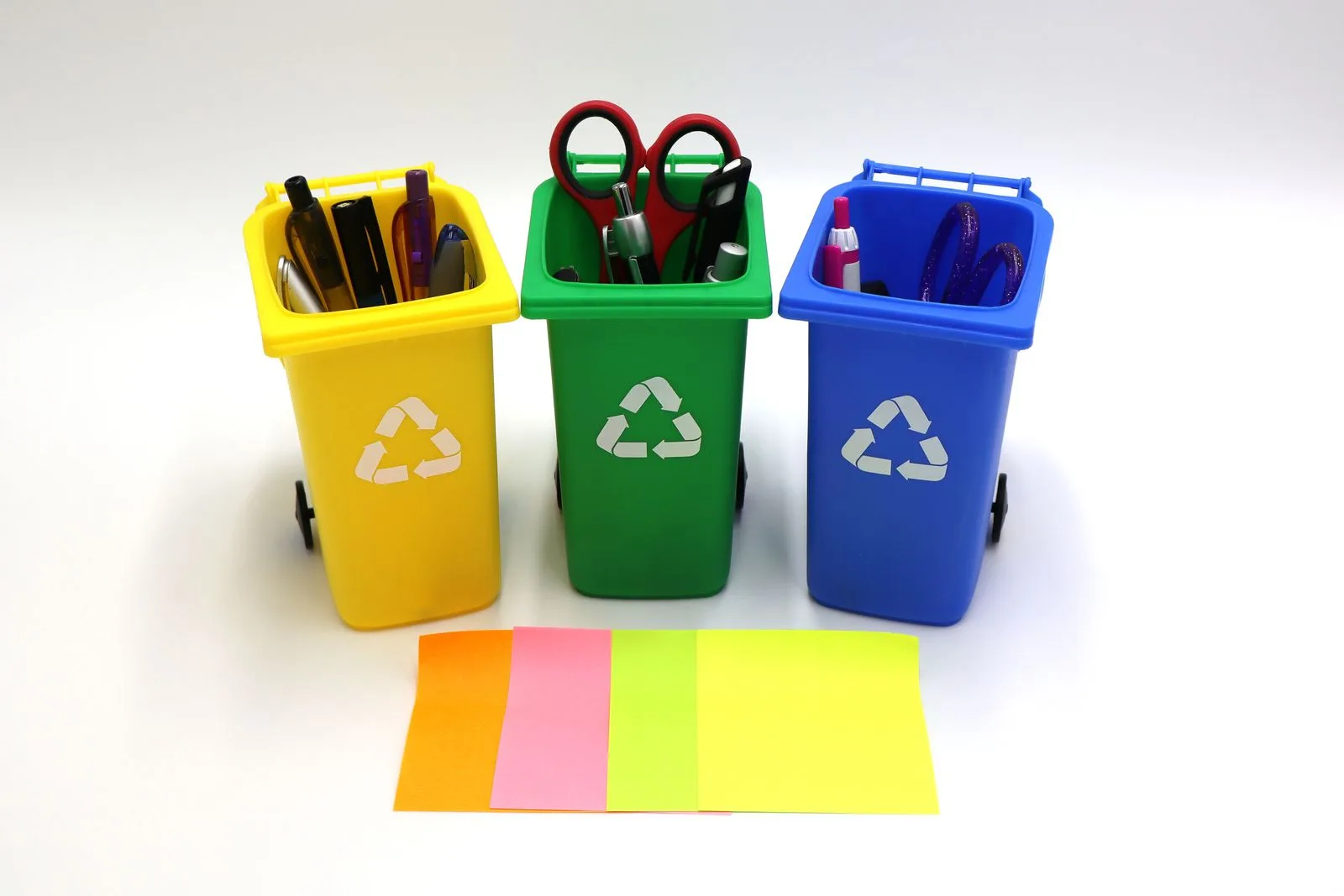 Mülltrennung im Büro: was zur Reinigung gehört und was nicht Drei farbige Mini Mülltonnen in Gelb, Grün und Blau mit Recyclingsymbol, gefüllt mit Büroutensilien, davor bunte Haftnotizen