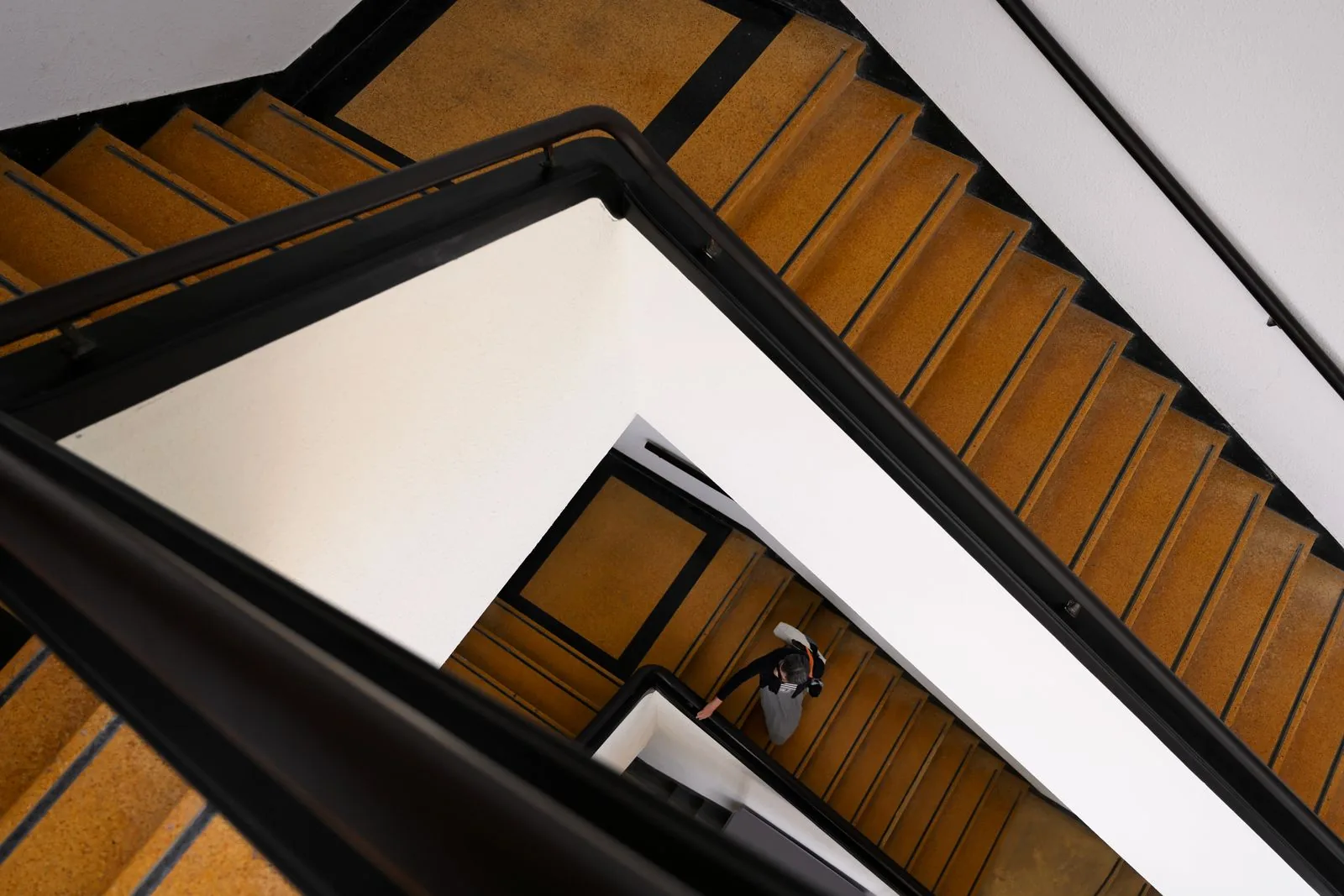 Putzplan im Treppenhaus: wann er funktioniert und wann nicht Blick von oben in ein mehrstöckiges Treppenhaus mit Terrazzostufen und schwarzem Handlauf, eine Person geht die Treppe hinunter
