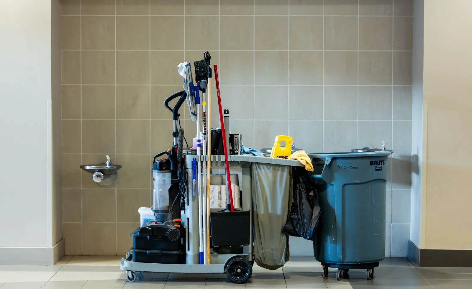 Eigene Reinigungskraft oder Reinigungsfirma: was sich rechnet Reinigungswagen mit Eimer, Mopp und Müllsack in einem gefliesten Flur