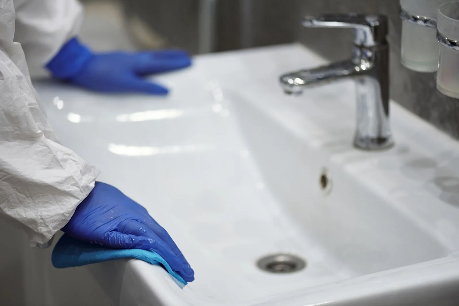 Sanitärreinigung im Büro: Reihenfolge und Fehlerquellen Hände in blauen Einmalhandschuhen wischen mit einem Tuch den Rand eines weißen Waschbeckens mit Chromarmatur