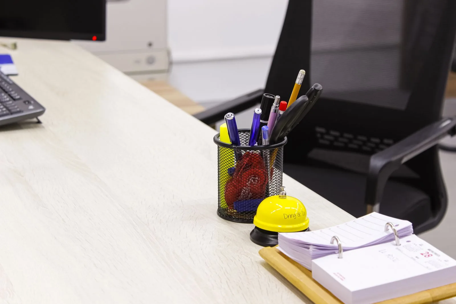 Schreibtisch reinigen im Büro: was die Reinigungsfirma übernimmt Heller Büroschreibtisch mit Tastatur, Stiftebecher, Tischklingel und Tischkalender vor einem schwarzen Bürostuhl