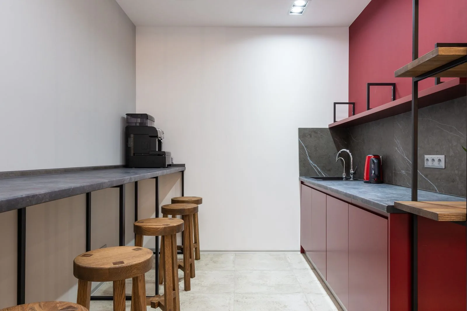 Teeküche im Büro: die drei häufigsten Hygienefallen Schmale Büroküche mit Kaffeevollautomat auf einer langen Theke, Holzhockern, Spüle und Wasserkocher vor roter Wand