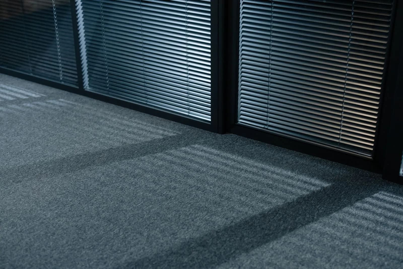 Teppichboden im Büro reinigen: Saugen, Flecken, Tiefenreinigung Nahaufnahme von grauem Teppichboden in einem hellen Großraumbüro
