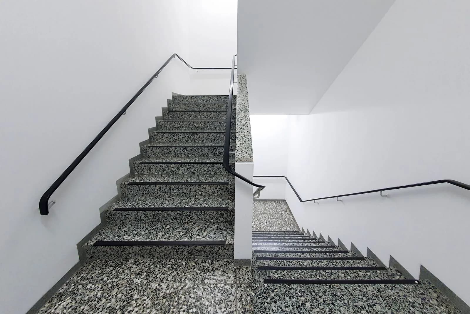 Was kostet die Treppenhausreinigung im Monat? Sauber gereinigtes Treppenhaus mit heller Granittreppe, schwarzem Handlauf und weißen Wänden