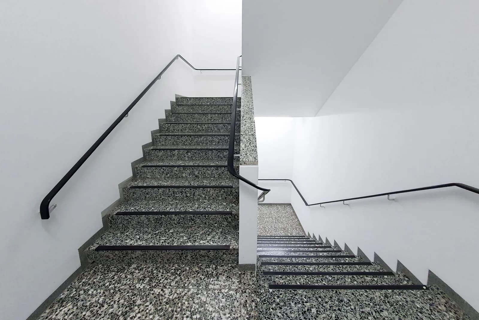 Treppenhausreinigung durch Mieter: wann die Pflicht gilt Helles Treppenhaus mit Terrazzostufen und schwarzem Handlauf an der weißen Wand
