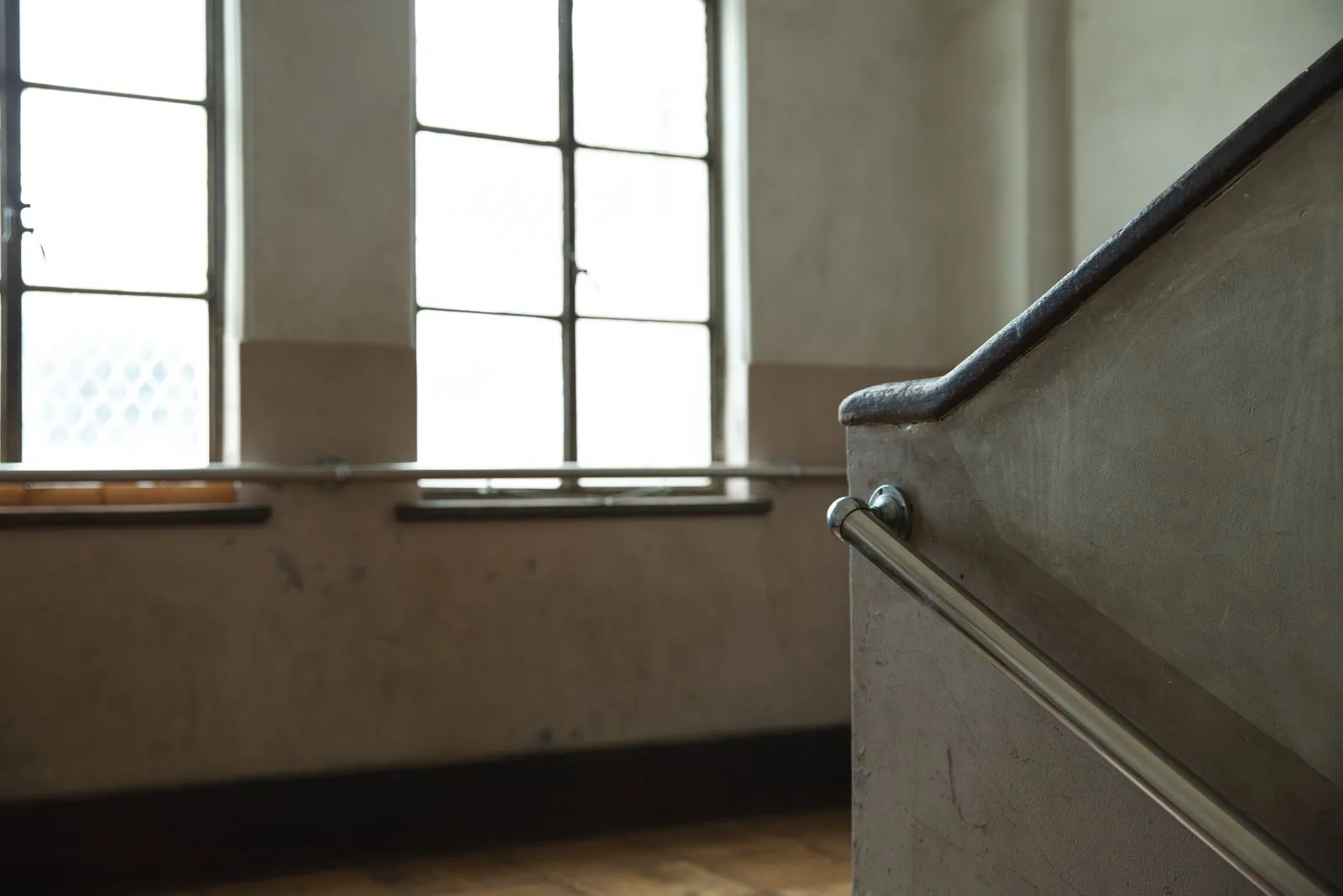 Was gehört zur Treppenhausreinigung? Der Leistungsumfang Treppenaufgang mit Edelstahlhandlauf an einer grauen Wand, dahinter zwei große Sprossenfenster mit Fensterbänken im Gegenlicht