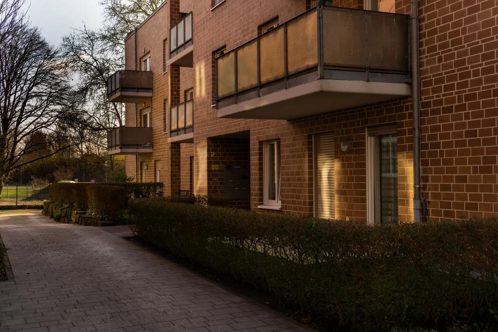 Treppenhausreinigung in der WEG: wer entscheidet und wer zahlt Wohnanlage mit roter Klinkerfassade, Balkonen und Hauseingang, davor ein gepflasterter Weg und eine niedrige Hecke im Abendlicht
